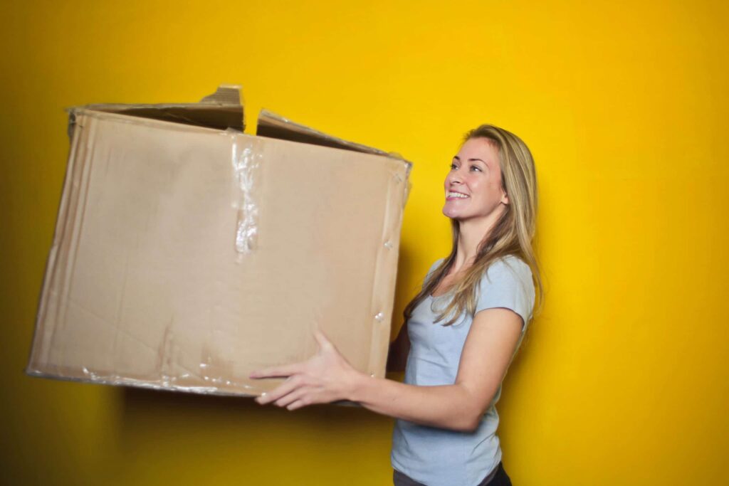 femme blonde portant un t-shirt bleu portant un gros cartons devant un fond jaune
